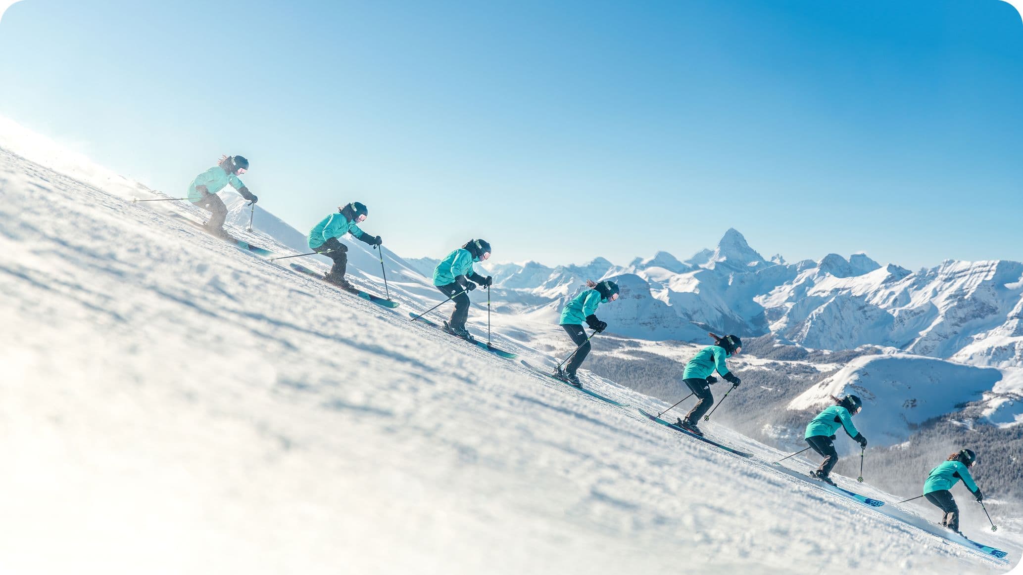 A skier in a turquoise jacket descends a snowy slope in a sequence of movements, with snow-capped mountains in the background under a clear blue sky.