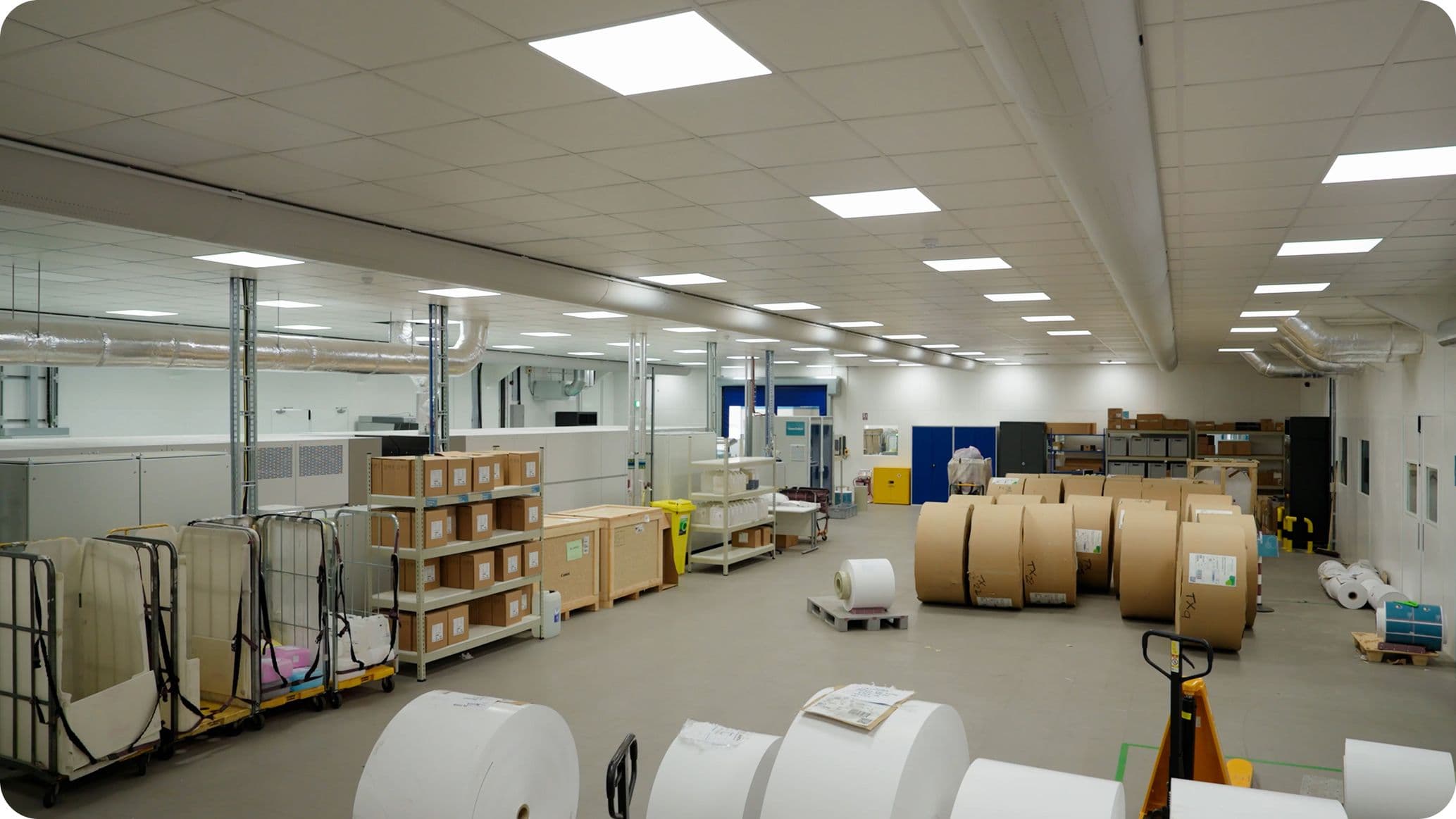 A spacious warehouse interior with large rolls of paper, shelves with boxes, and industrial equipment under bright fluorescent lighting.