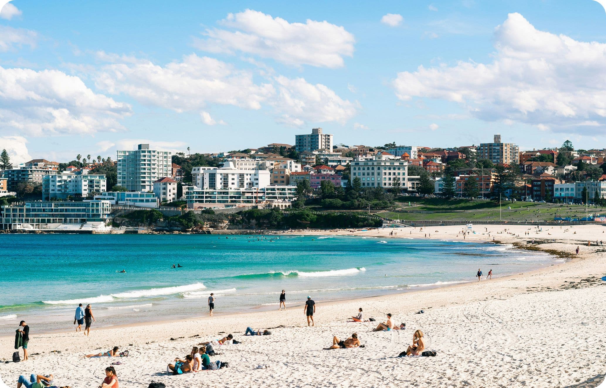 Sandy beach with people sunbathing and swimming, turquoise water, and city buildings in the background under a partly cloudy sky.