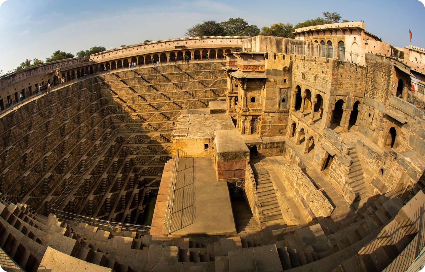 A wide-angle view of an ancient stepwell with intricate stone steps and arches, surrounded by historic architecture under a clear sky.