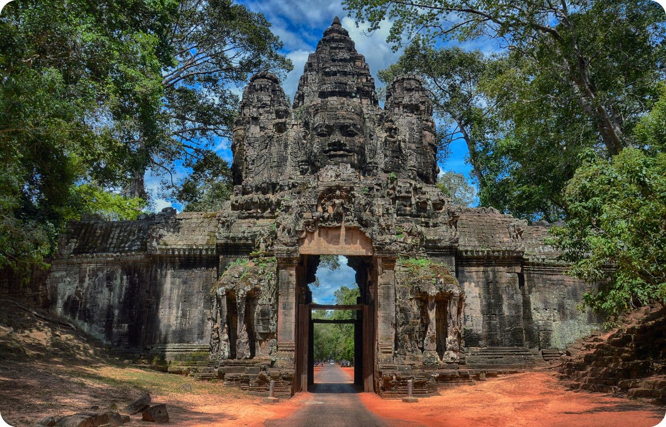 Ancient stone gate with carved faces at Angkor Thom, Cambodia, surrounded by lush greenery under a blue sky.