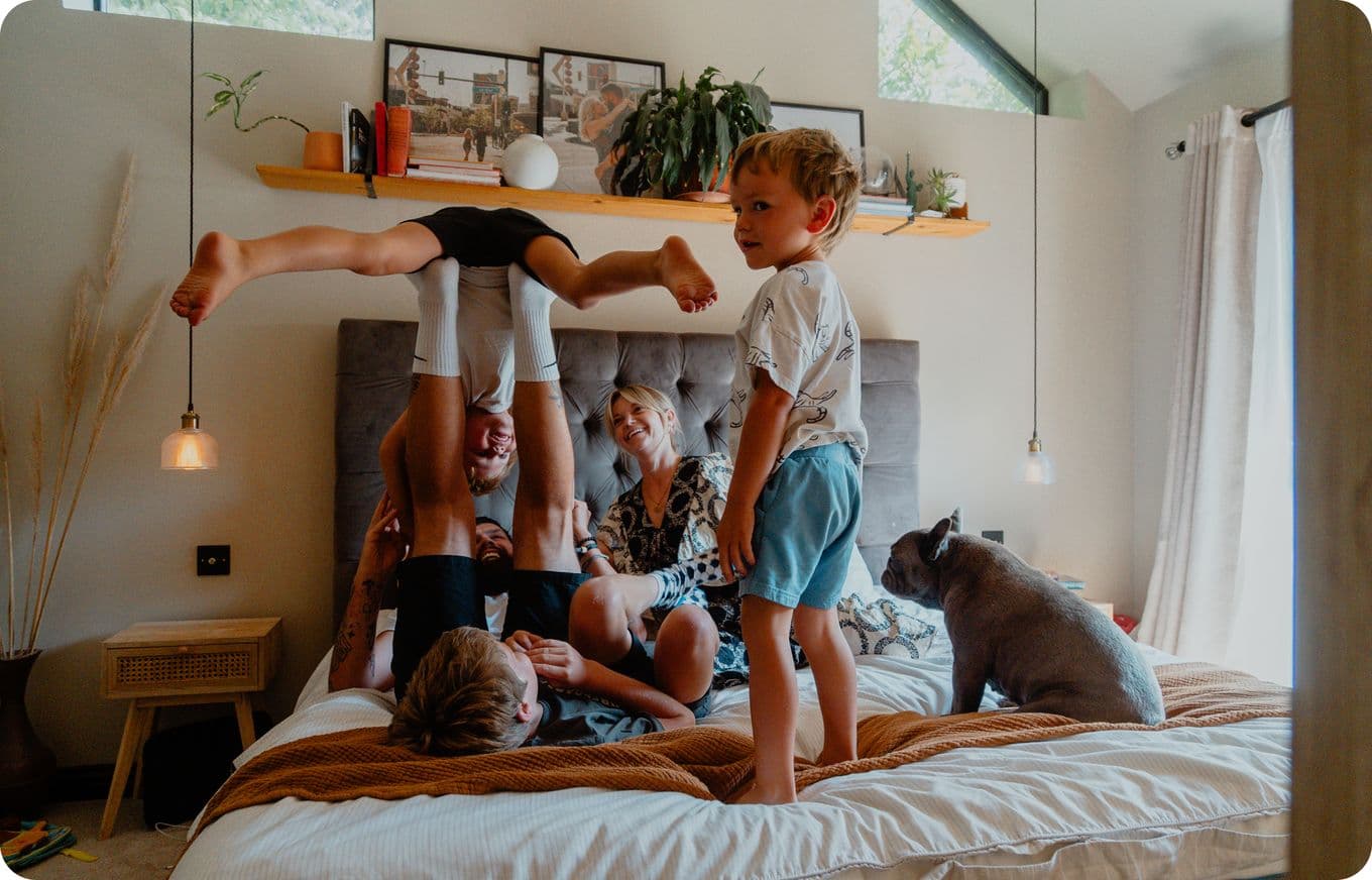 A joyful family plays on a bed with a dog nearby; one child is upside down, and others are laughing. Cozy room with shelves and decor.