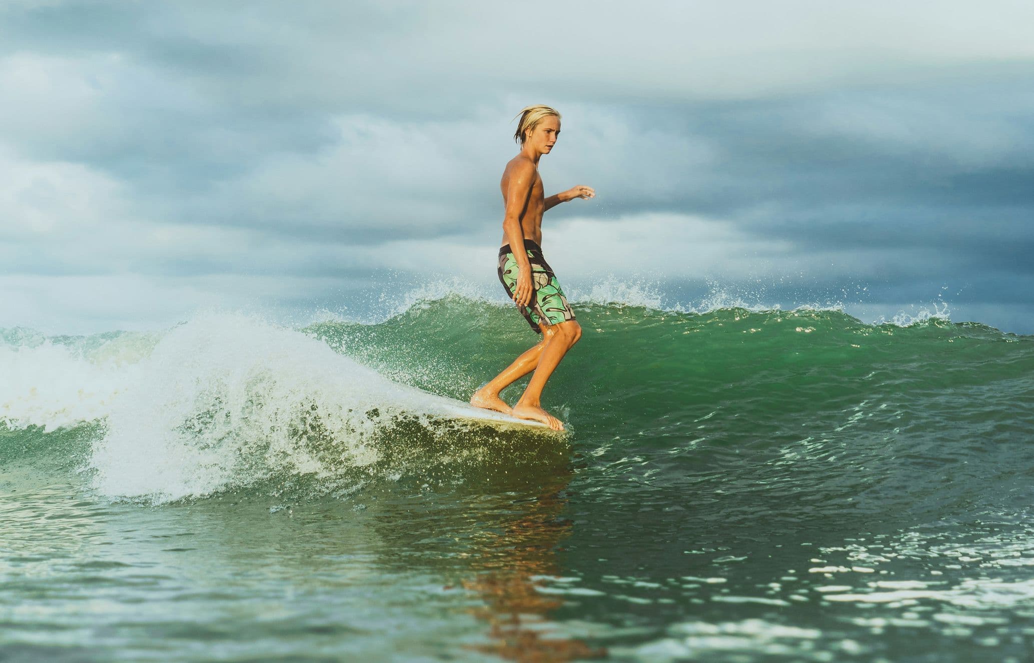 Shirtless blond surfer in patterned shorts rides a longboard on a green wave beneath a cloudy sky.