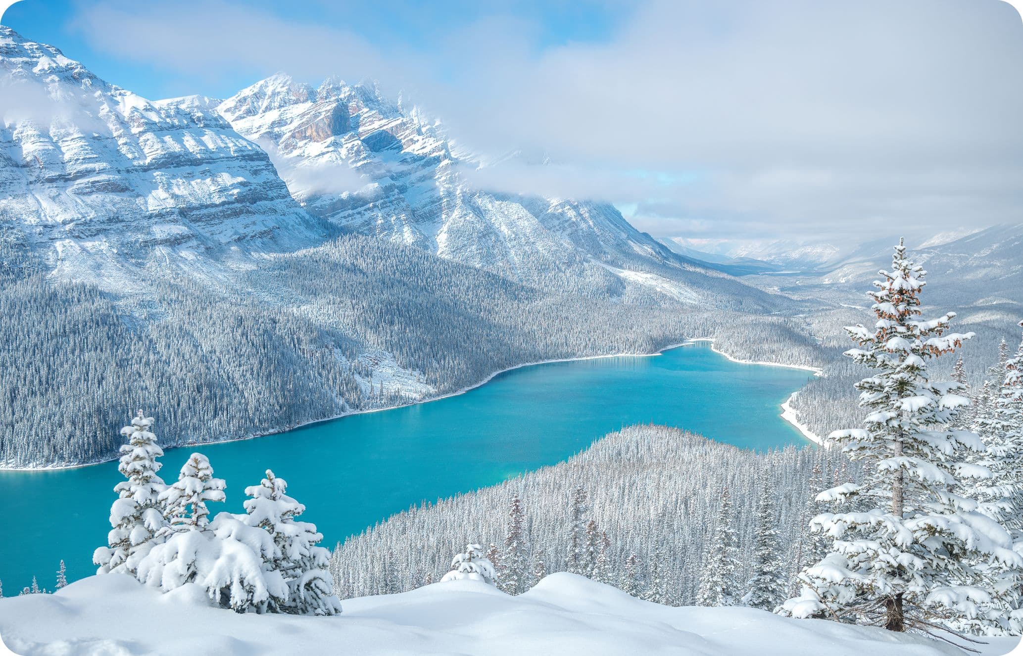 Snow-covered mountains and trees surround a turquoise lake under a partly cloudy sky, creating a serene winter landscape.