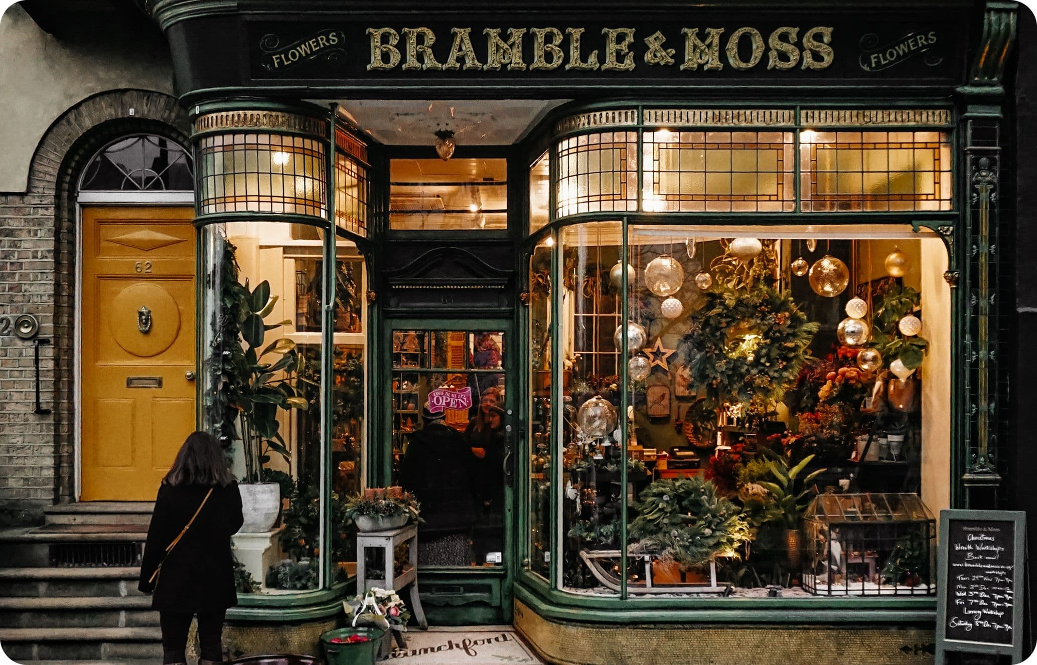 A quaint flower shop named "Bramble & Moss" with a green facade, large display windows, and a yellow door. A person stands outside admiring the decor.