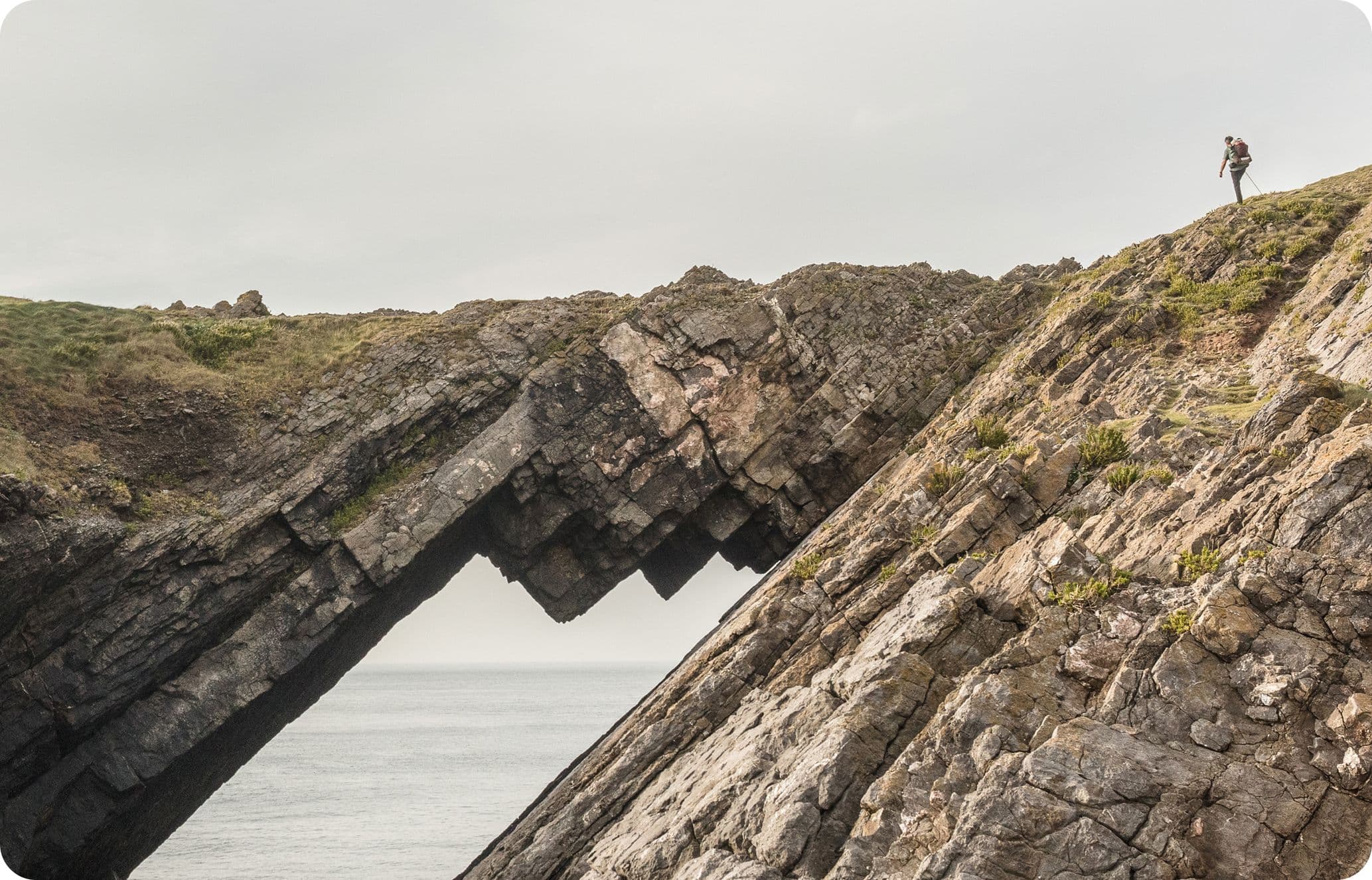 A person walks on a rocky, arch-shaped cliff above the ocean under a cloudy sky.