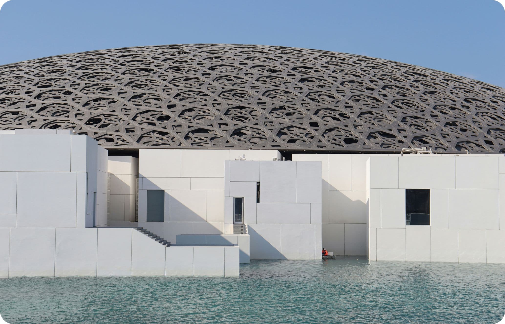 Modern building with intricate dome and white geometric walls, surrounded by water under a clear blue sky.