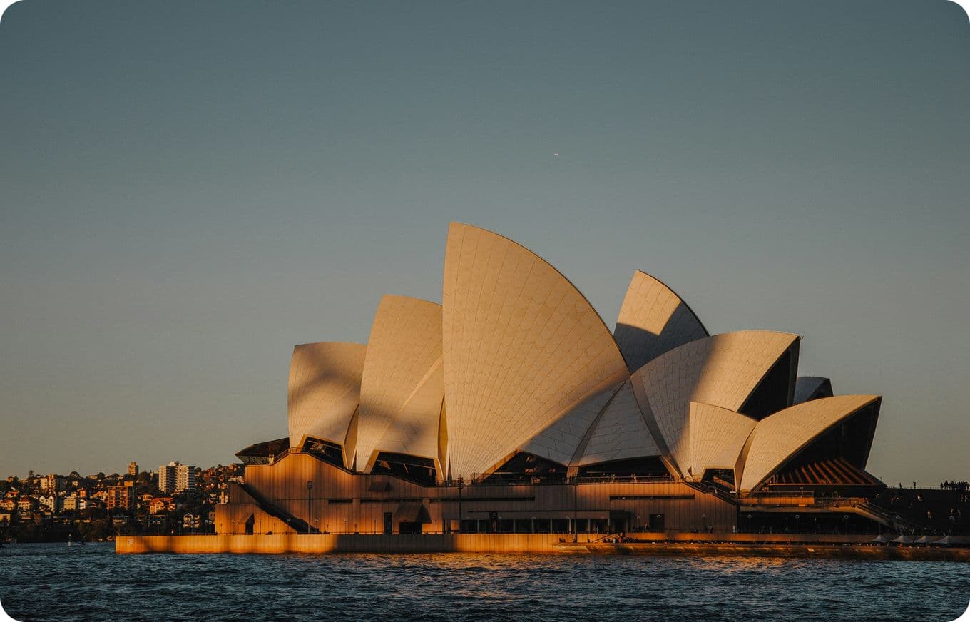 Sydney Opera House illuminated by golden sunset light, with a clear sky and water in the foreground.