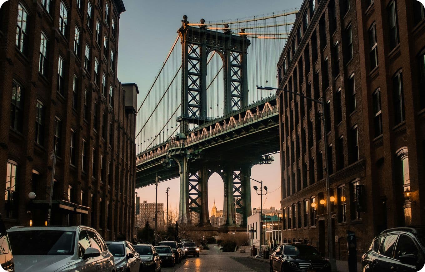 View of a bridge framed by tall brick buildings at sunset, with parked cars lining the street below.
