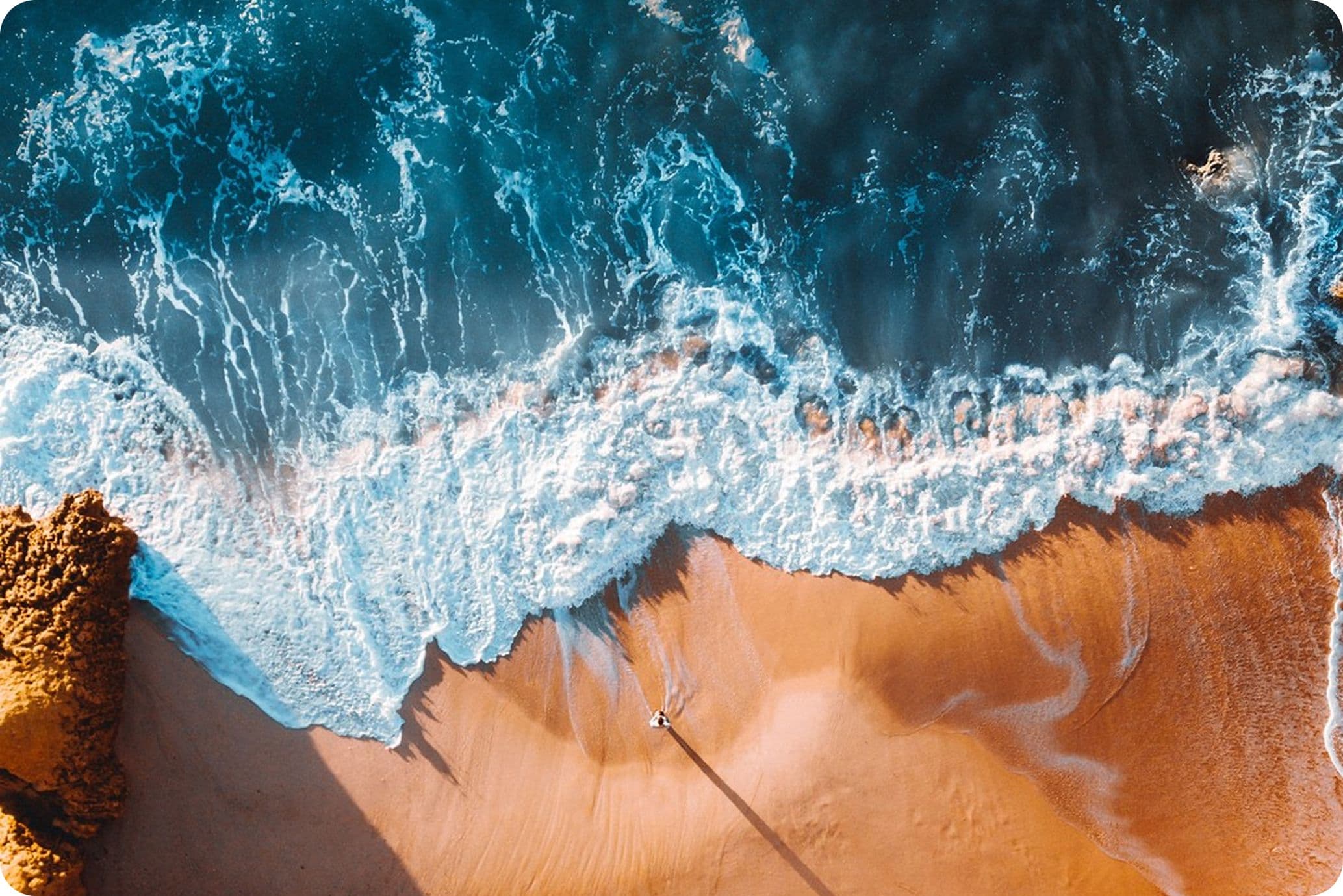 Aerial view of waves crashing onto a sandy beach, with a person casting a long shadow, and rocky formations on the left.