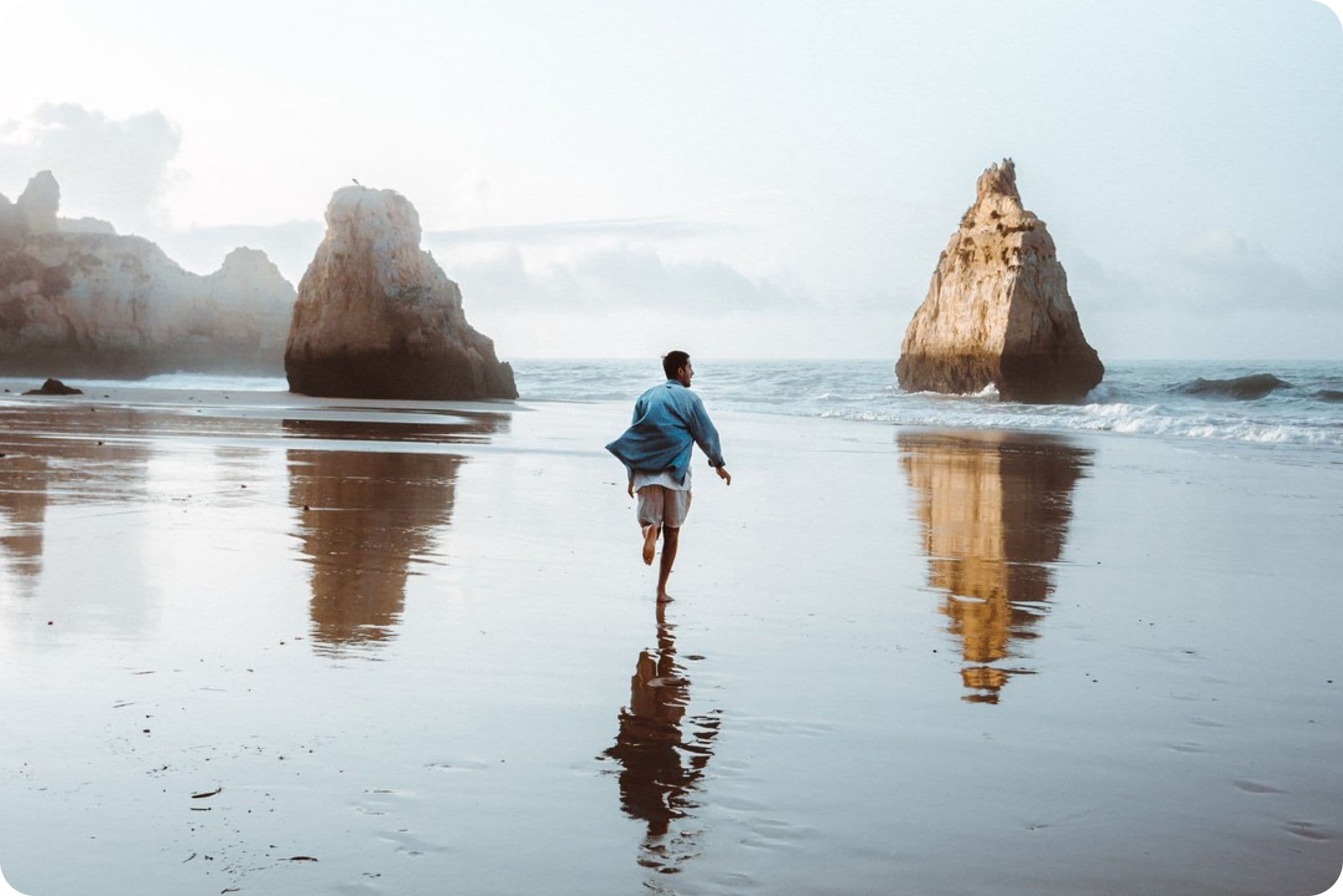 Person running on a reflective beach towards large rock formations in the sea, under a cloudy sky.