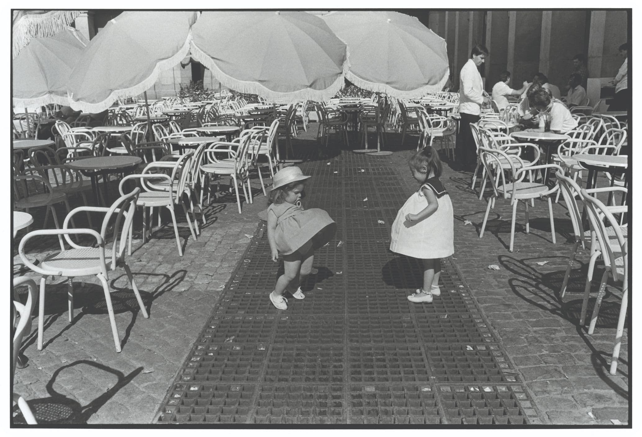 Black-and-white photo of two small children, one in a hat crouching and the other standing, amid empty café tables, chairs and large umbrellas.