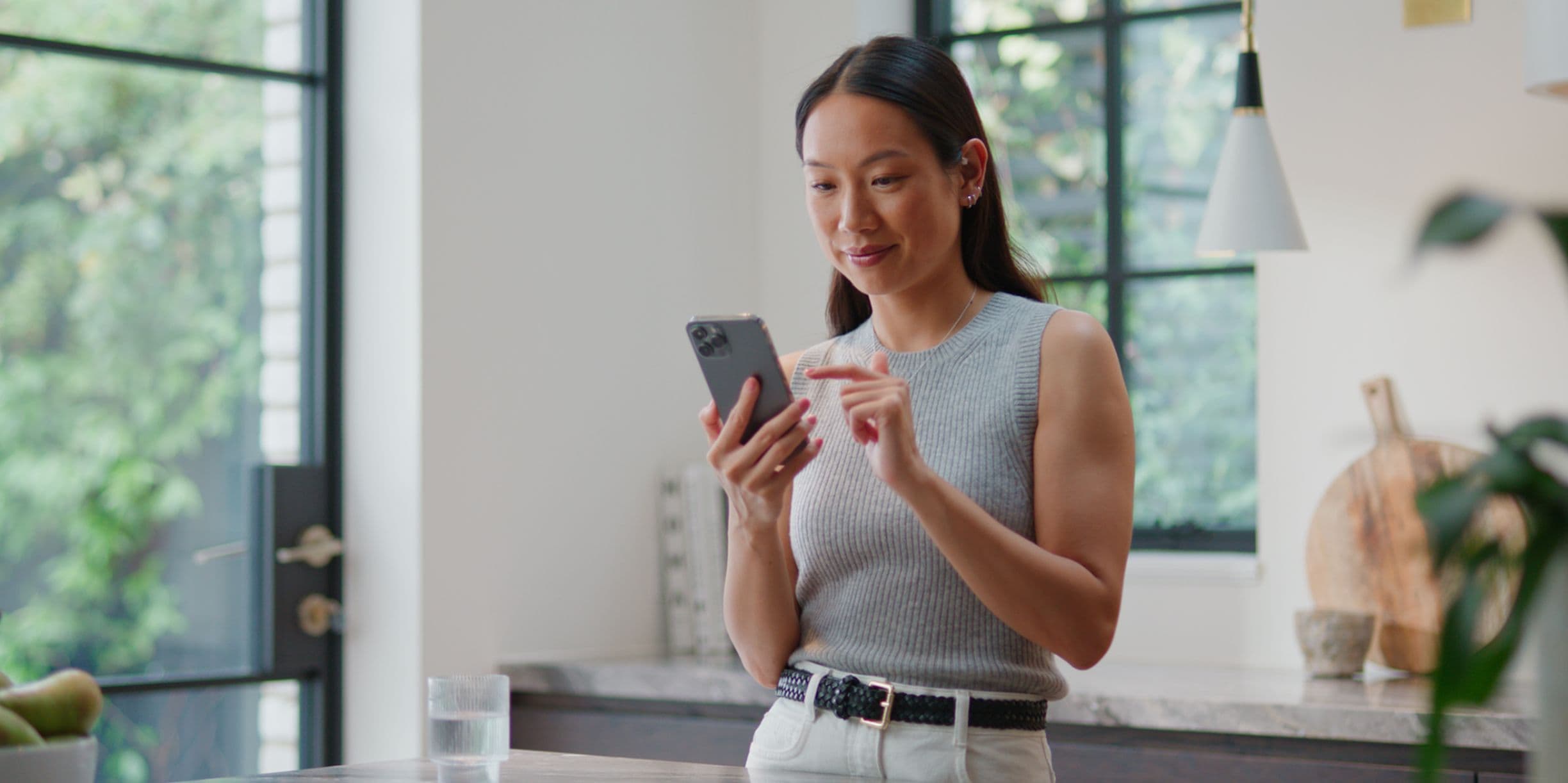 Woman in a kitchen, wearing a gray sleeveless top, smiles while using a smartphone. A glass of water is on the counter.