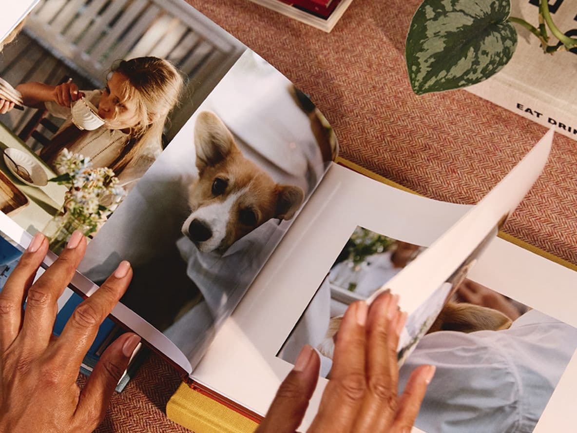Hands flipping through a photo book showing images of a woman drinking coffee and a dog. Nearby, a book titled "Eat Drink Nap."