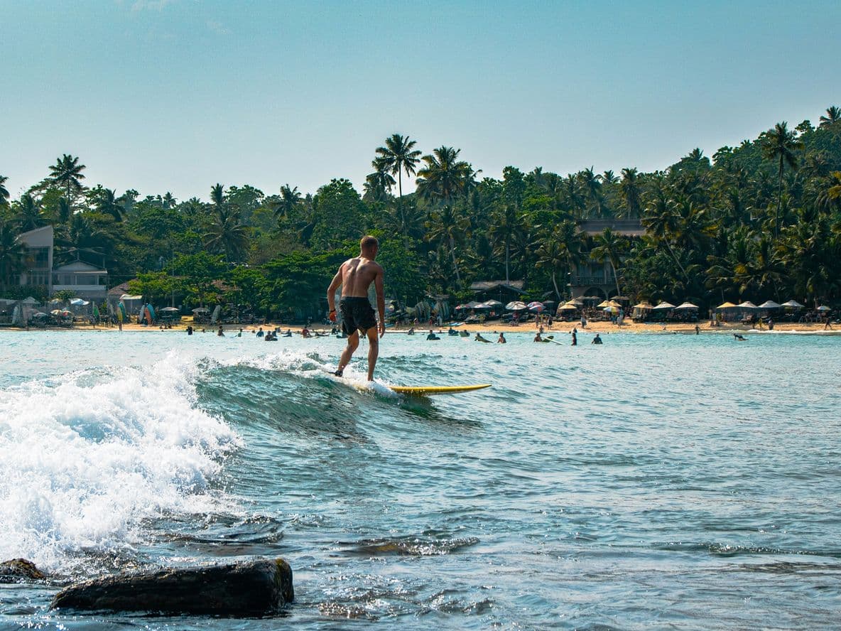 A person surfing on a small wave near a tropical beach with palm trees and distant beachgoers under umbrellas.