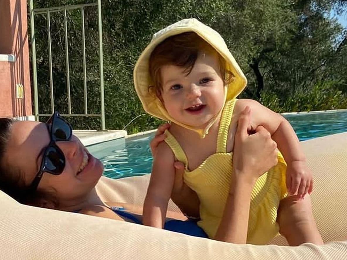 Woman lounging on a pool float, holding a smiling baby in a yellow outfit with a hood. Trees and a building are in the background.