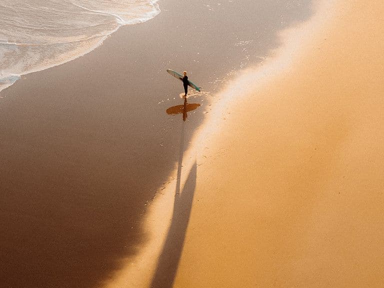 An aerial photogrpah of a surfer approaching the water from a sandy beach, shot from a drone