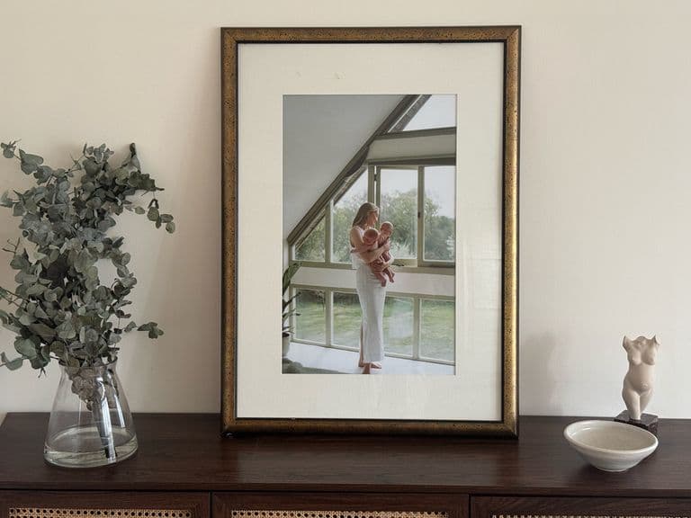 Framed photo on a wooden cabinet with a vase of greenery and a small bowl, against a plain wall.