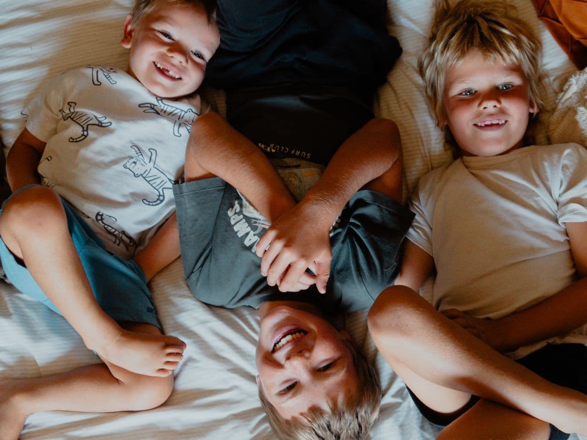Three children lying on a bed, smiling and playing together, with a dog resting nearby.