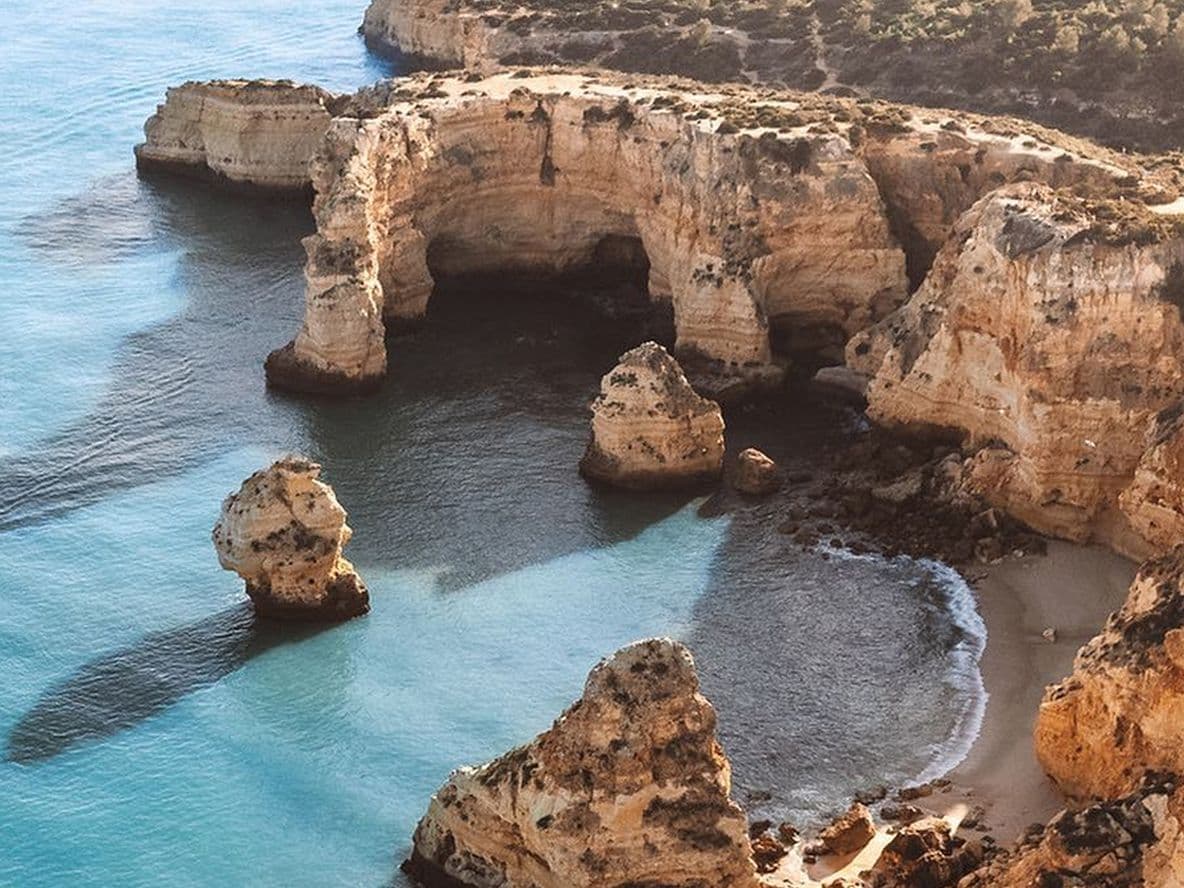 Aerial view of rocky cliffs and clear blue waters along a coastline, with a small boat leaving a wake near the shore.