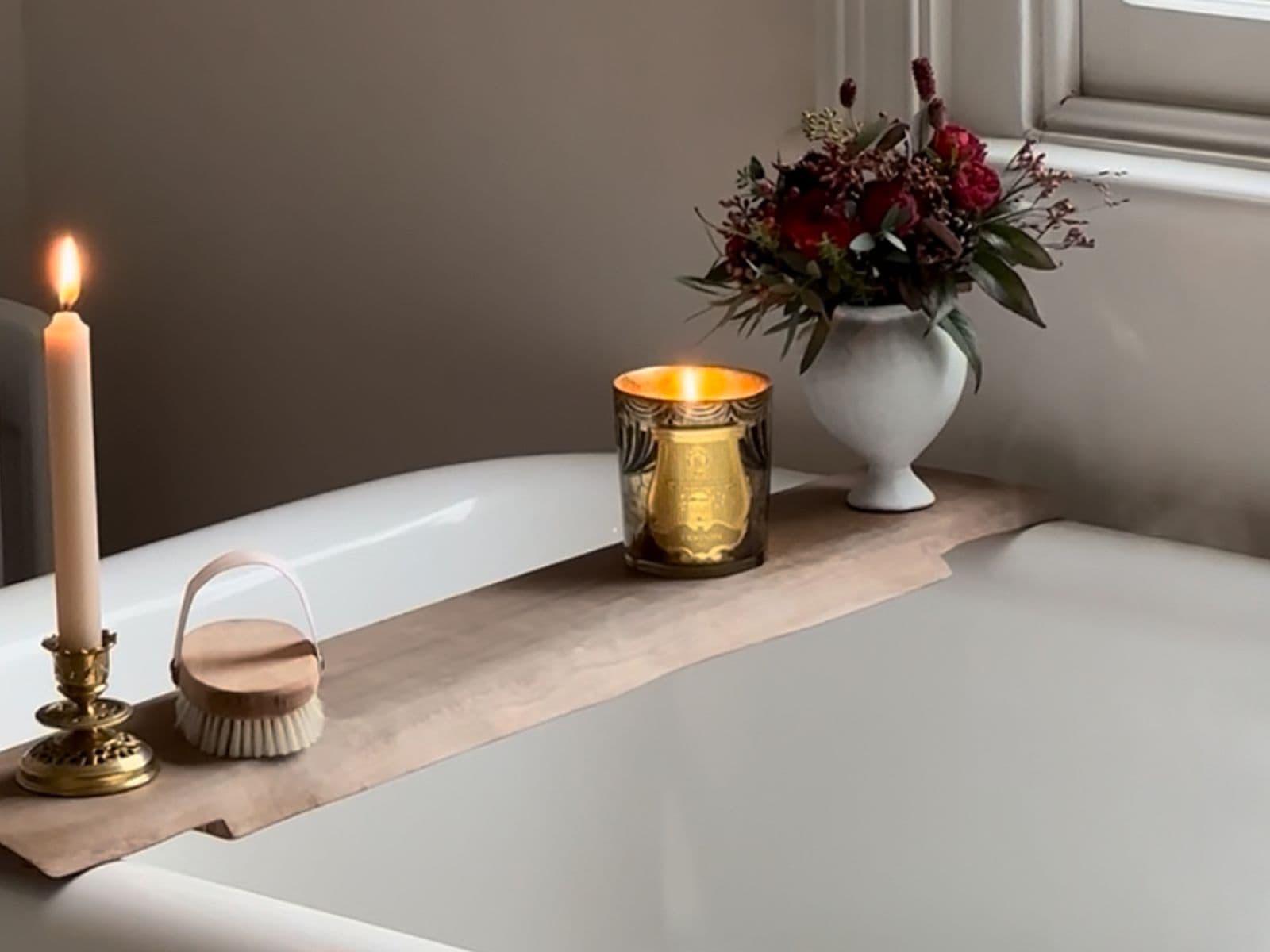 A lit candle, a gold candle holder, a brush, and flowers on a wooden board across a white bathtub with running water.