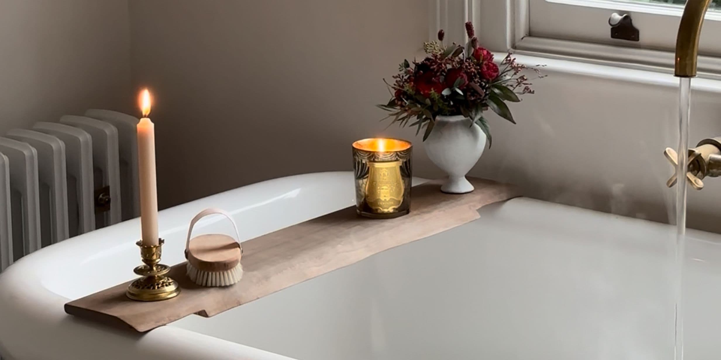 A lit candle, a gold candle holder, a brush, and flowers on a wooden board across a white bathtub with running water.