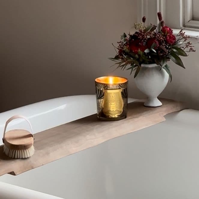 A lit candle, a gold candle holder, a brush, and flowers on a wooden board across a white bathtub with running water.