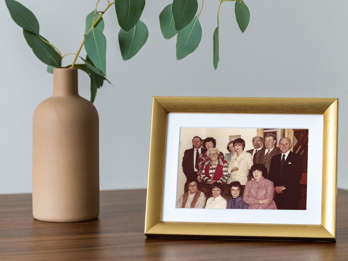 A framed family photo on a wooden table next to a beige vase with green leaves.