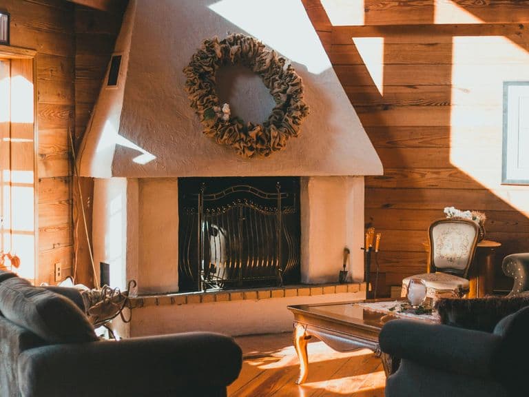 Cozy living room with a white fireplace, wreath, wooden walls, and sunlight streaming through large windows onto sofas and a coffee table.
