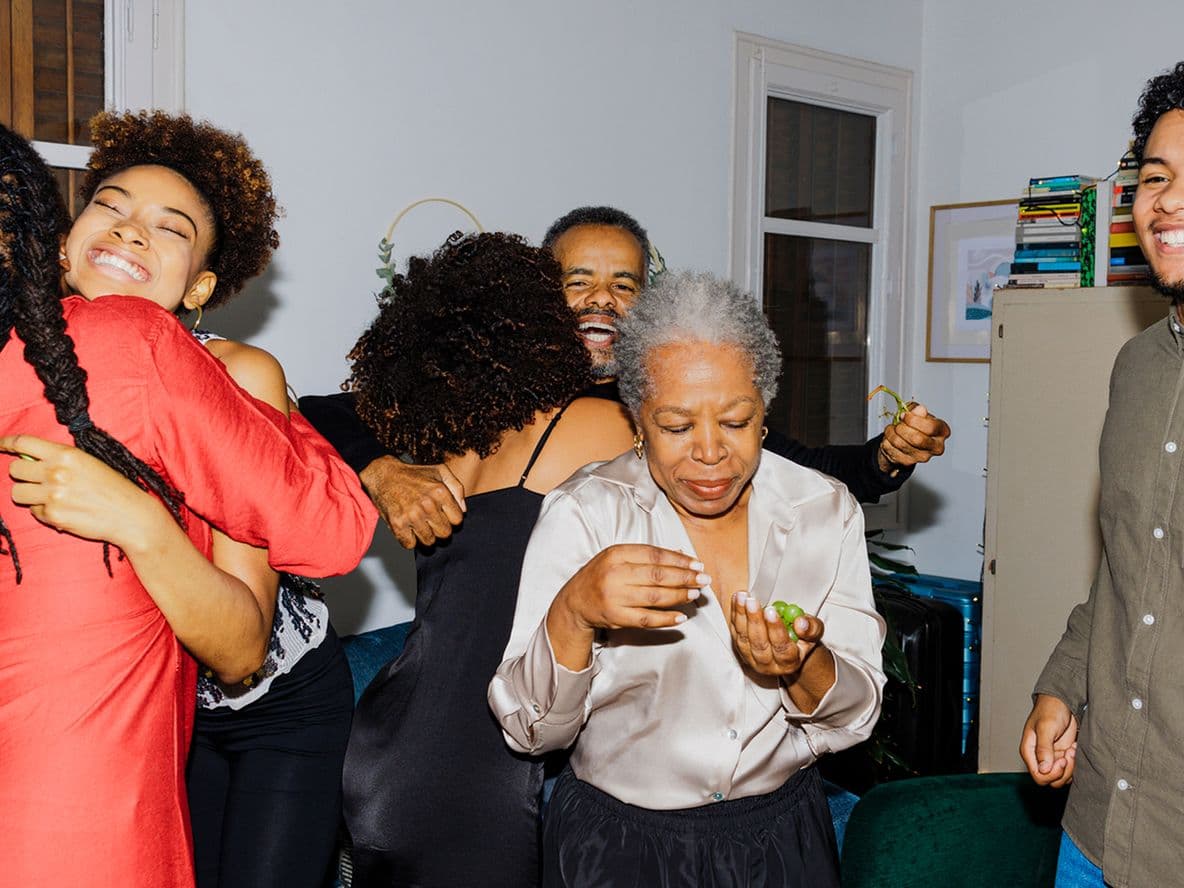 A joyful family gathering with people hugging and smiling in a small room, featuring a woman in a red dress and books on a shelf.