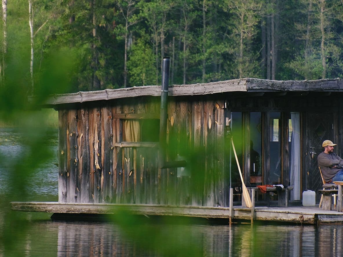 Man sitting on the deck of a rustic floating wooden cabin on a calm forest lake, blurred leaves in the foreground.