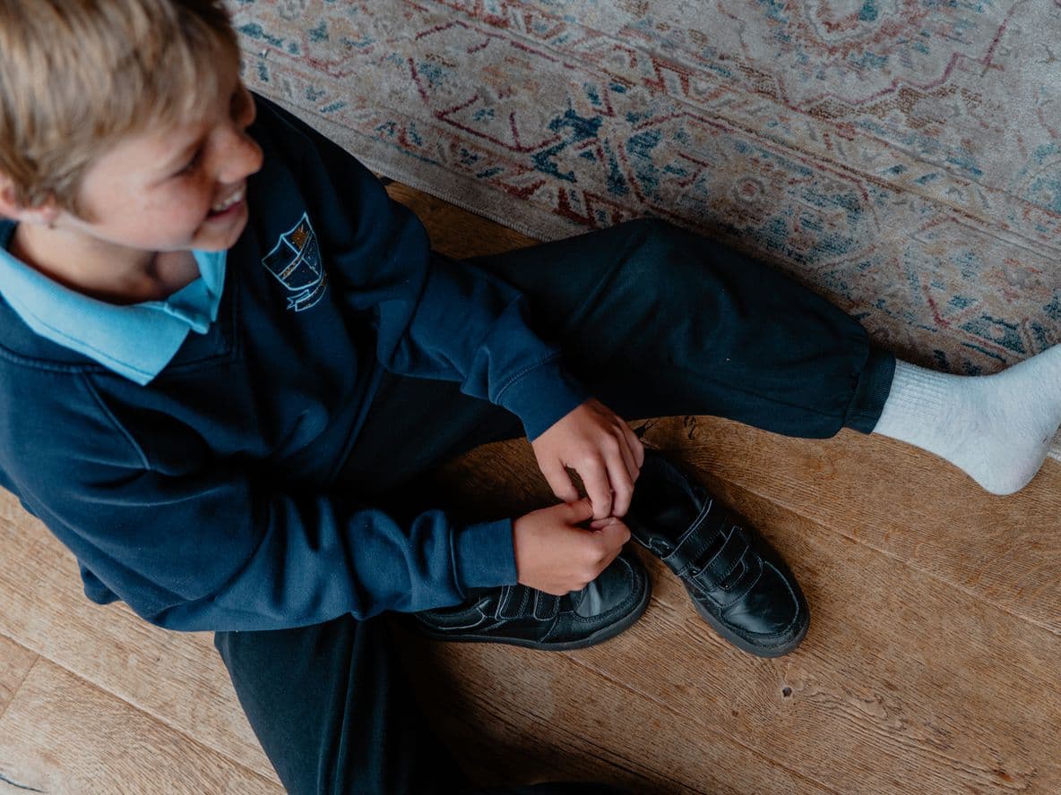 A child sits on a wooden floor, wearing a school uniform, tying the laces of one shoe while the other foot is covered in a white sock.