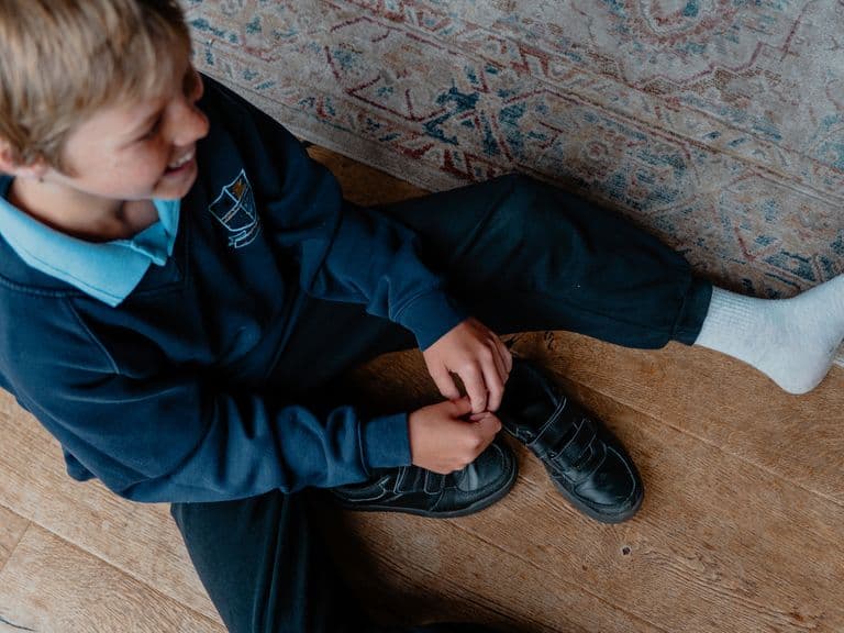 A child sits on a wooden floor, wearing a school uniform, tying the laces of one shoe while the other foot is covered in a white sock.