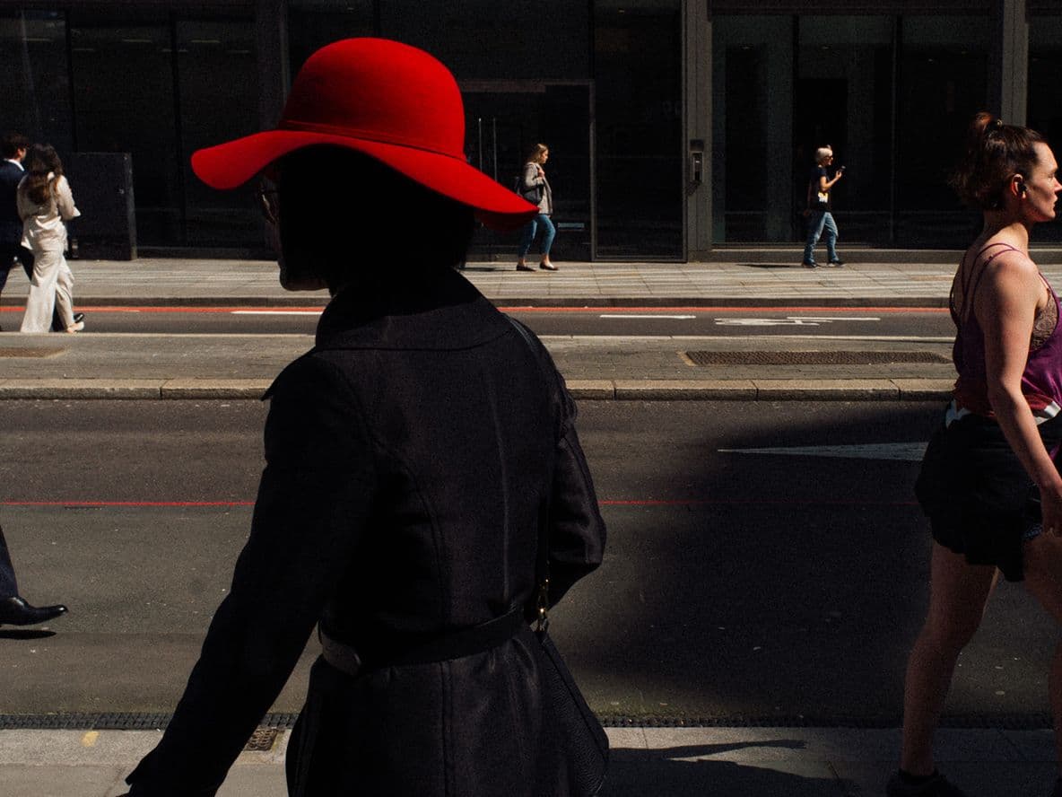 Silhouetted woman in a red hat walks on a city street, with people in the background against a modern building facade.