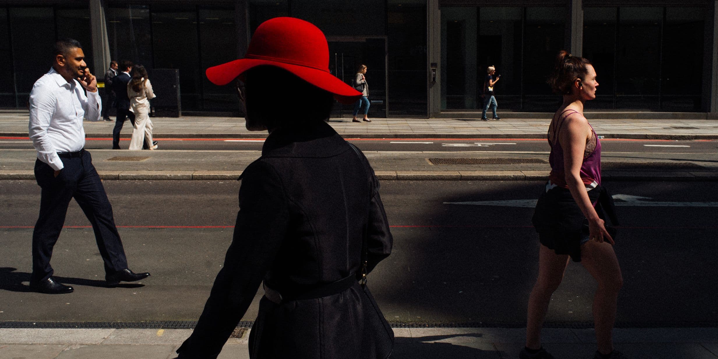 Silhouetted woman in a red hat walks on a city street, with people in the background against a modern building facade.