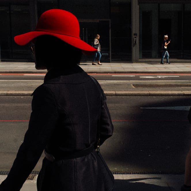 Silhouetted woman in a red hat walks on a city street, with people in the background against a modern building facade.