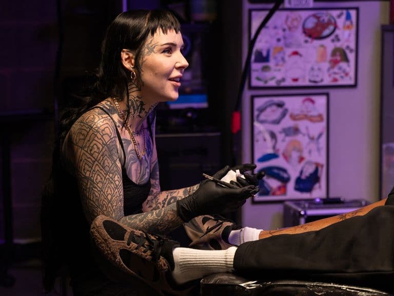 A tattoo artist with long dark hair and tattoos on her arms and face works on a client's leg in a dimly lit studio.
