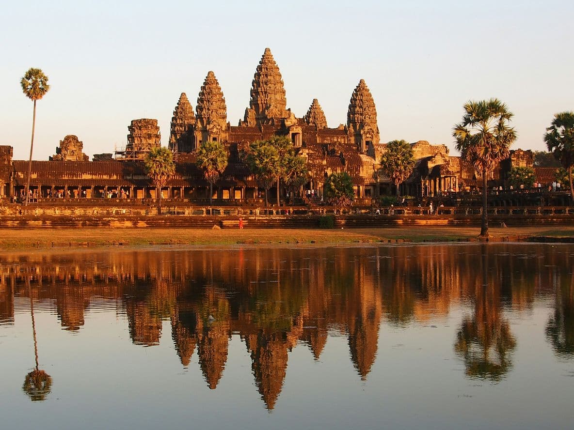 Angkor Wat temple complex at sunset, reflected in a calm pond, surrounded by palm trees and a clear sky.