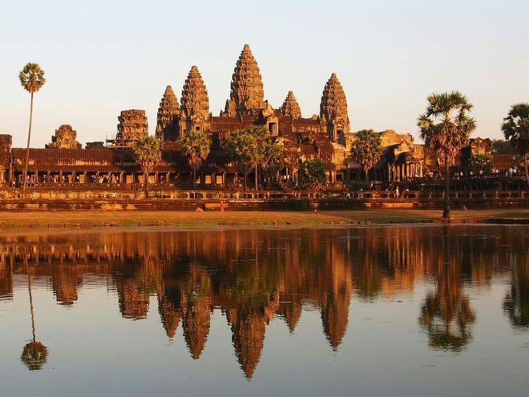 Angkor Wat temple complex at sunset, reflected in a calm pond, surrounded by palm trees and a clear sky.