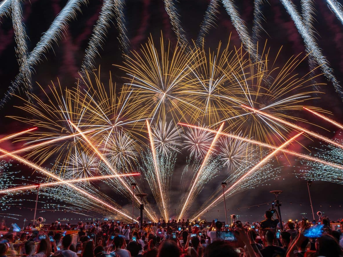 A vibrant fireworks display lights up the night sky, with a crowd of people watching and capturing the moment on their phones.
