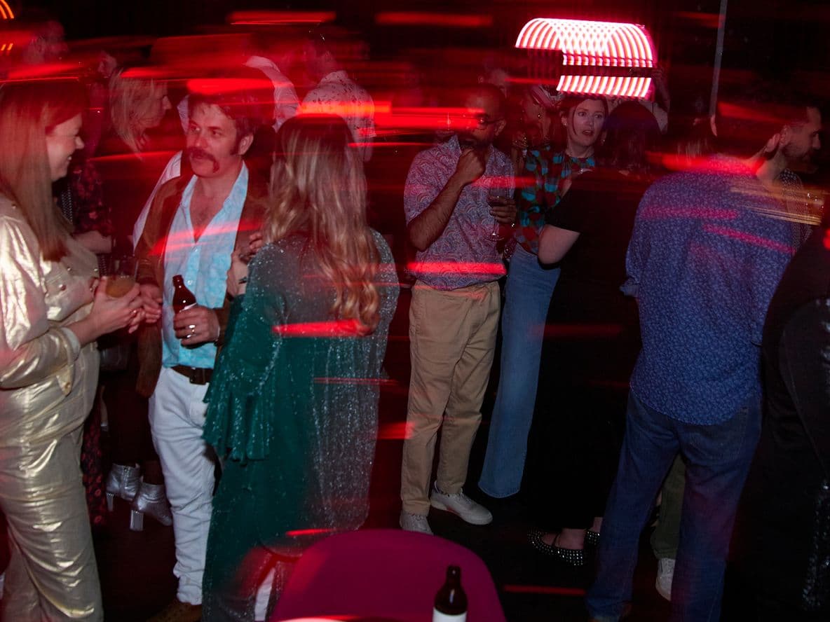 People at a lively party with red light streaks, wearing casual and semi-formal attire, some holding drinks, chatting, and mingling.