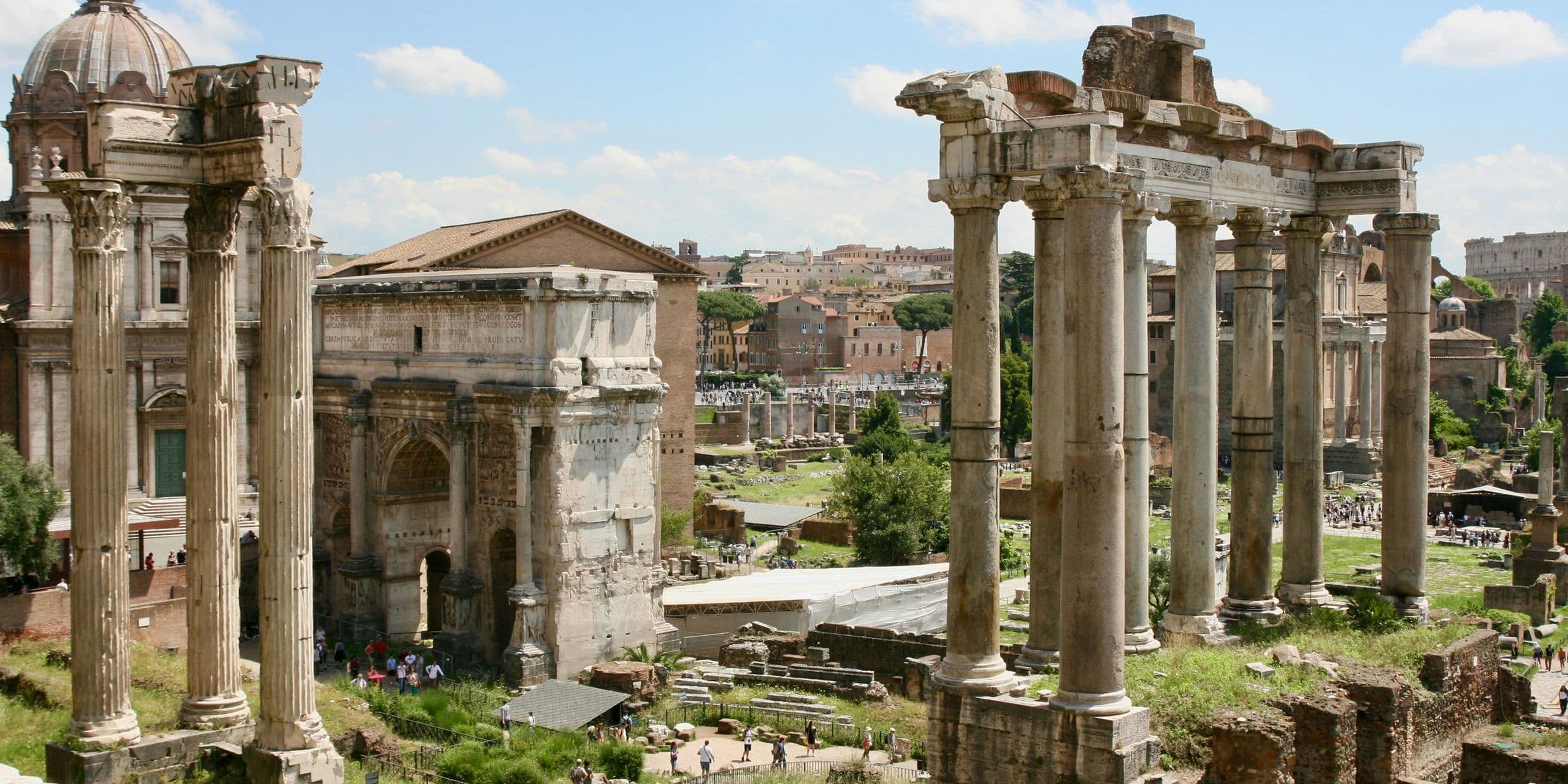 Ancient Roman ruins with tall columns and historic buildings under a blue sky with scattered clouds.