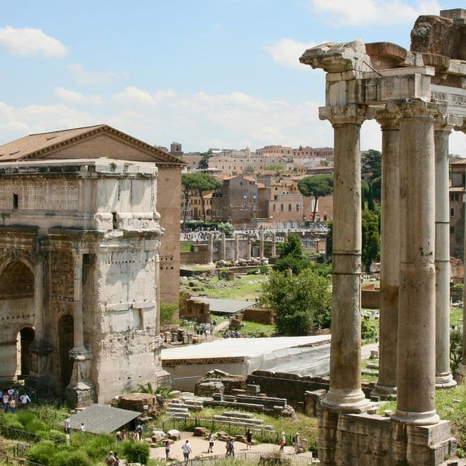 Ancient Roman ruins with tall columns and historic buildings under a blue sky with scattered clouds.