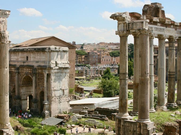 Ancient Roman ruins with tall columns and historic buildings under a blue sky with scattered clouds.
