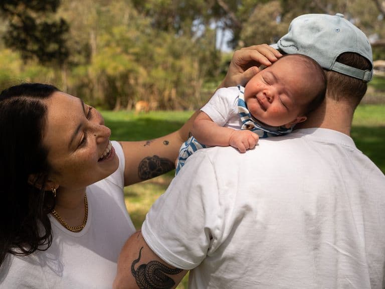 A woman smiles at a baby resting on a man's shoulder outdoors in a park. The baby is wearing a patterned outfit and a cap.