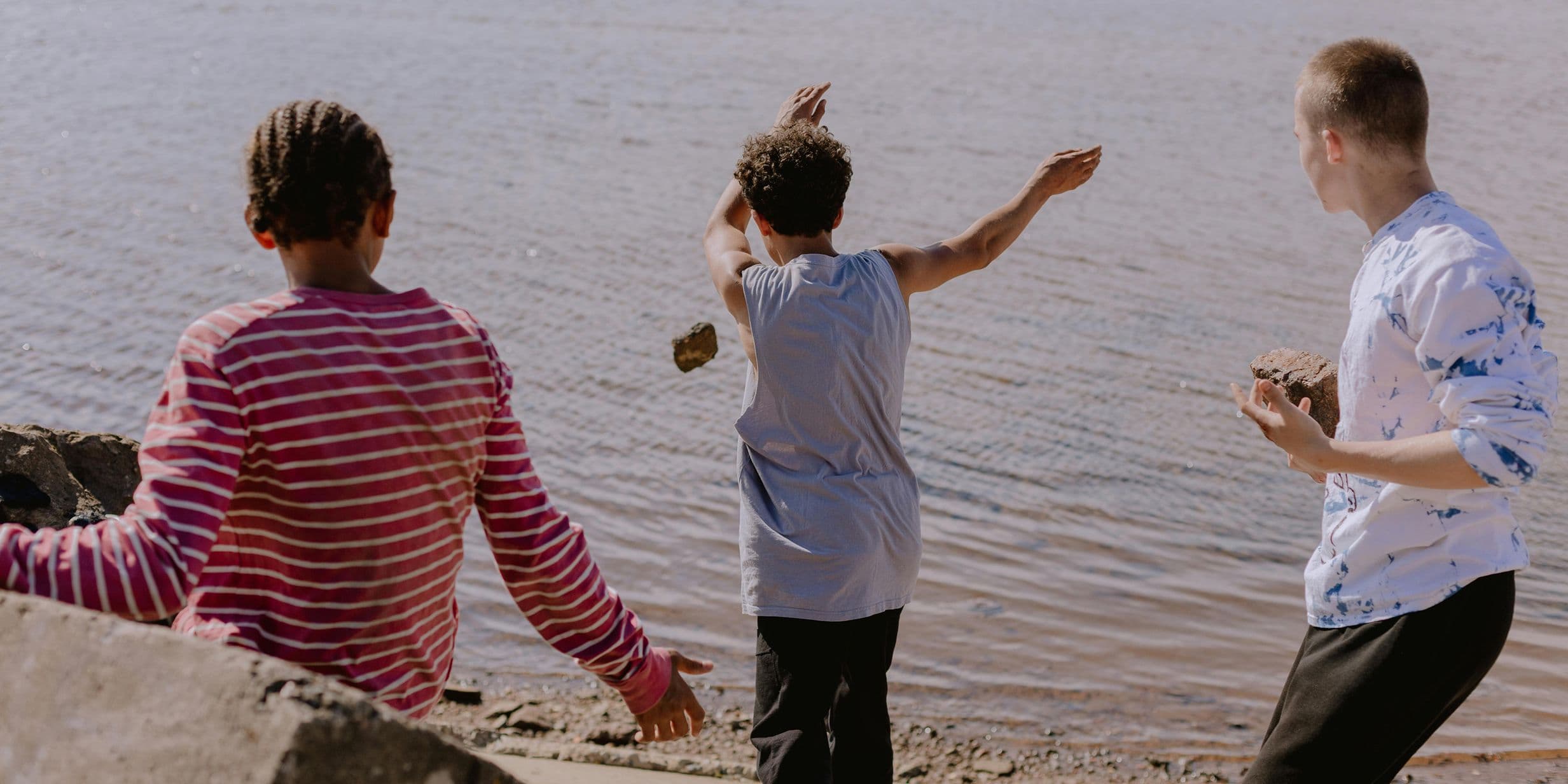 Three boys on a lakeshore skipping stones; center boy mid-throw, others watching from the shore.