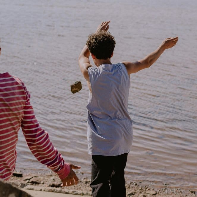 Three boys on a lakeshore skipping stones; center boy mid-throw, others watching from the shore.