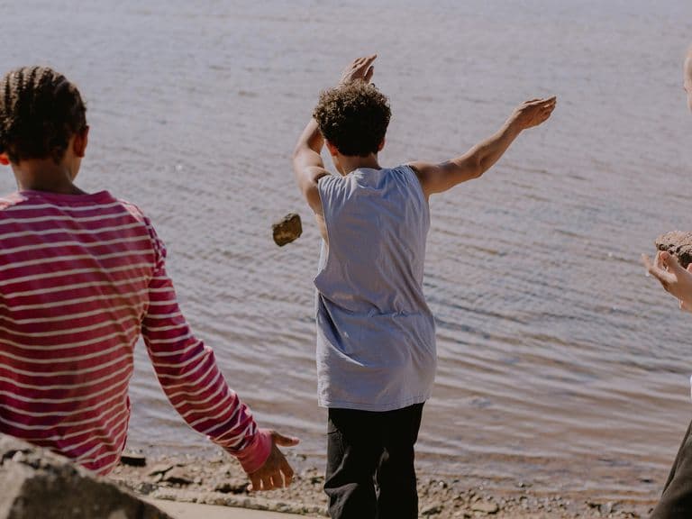 Three boys on a lakeshore skipping stones; center boy mid-throw, others watching from the shore.