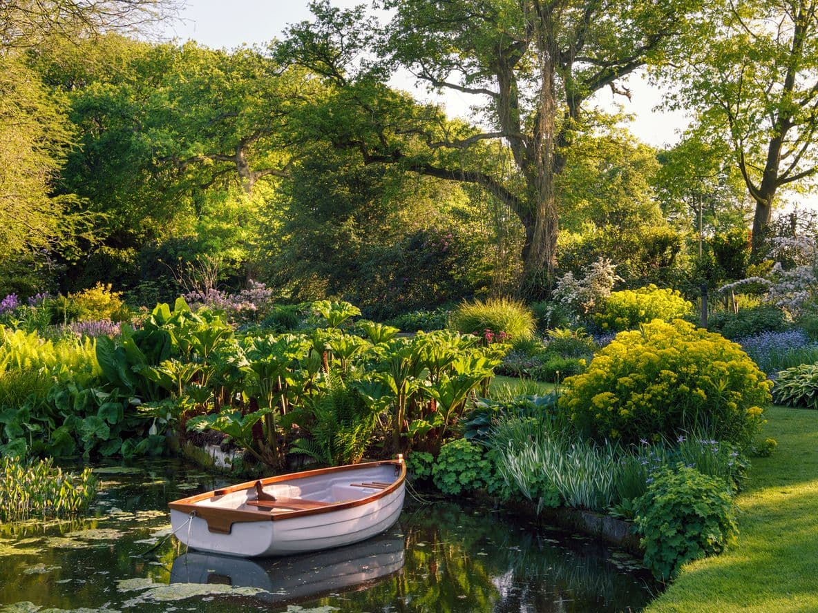 A small wooden boat floats on a tranquil pond surrounded by lush greenery and colorful flowers in a sunlit garden.