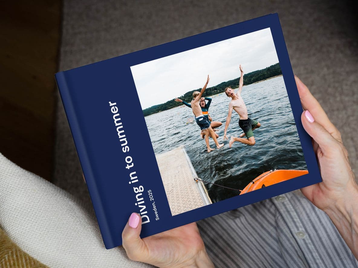 Person holding a photo book titled "Diving in to summer," featuring an image of two people jumping into a lake from a dock.